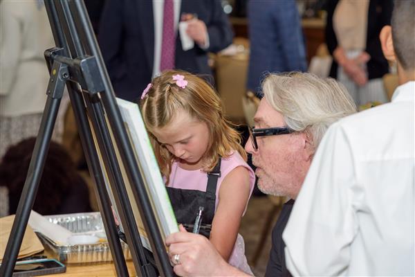 An artist crouches down beside a young student. Together, they are working on painting a work of art. 